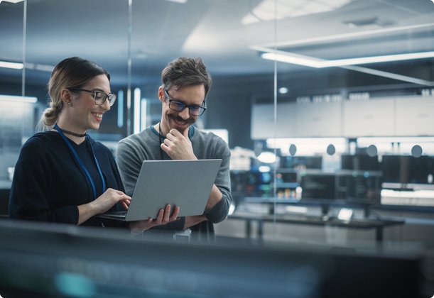 Two smiling business people, a man and a woman with glasses, stand in a modern office and look together at a laptop.