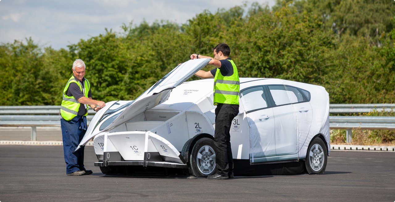 Two technicians in yellow safety vests are working on a white test vehicle, which is partially covered or modular in design, on an outdoor test track.