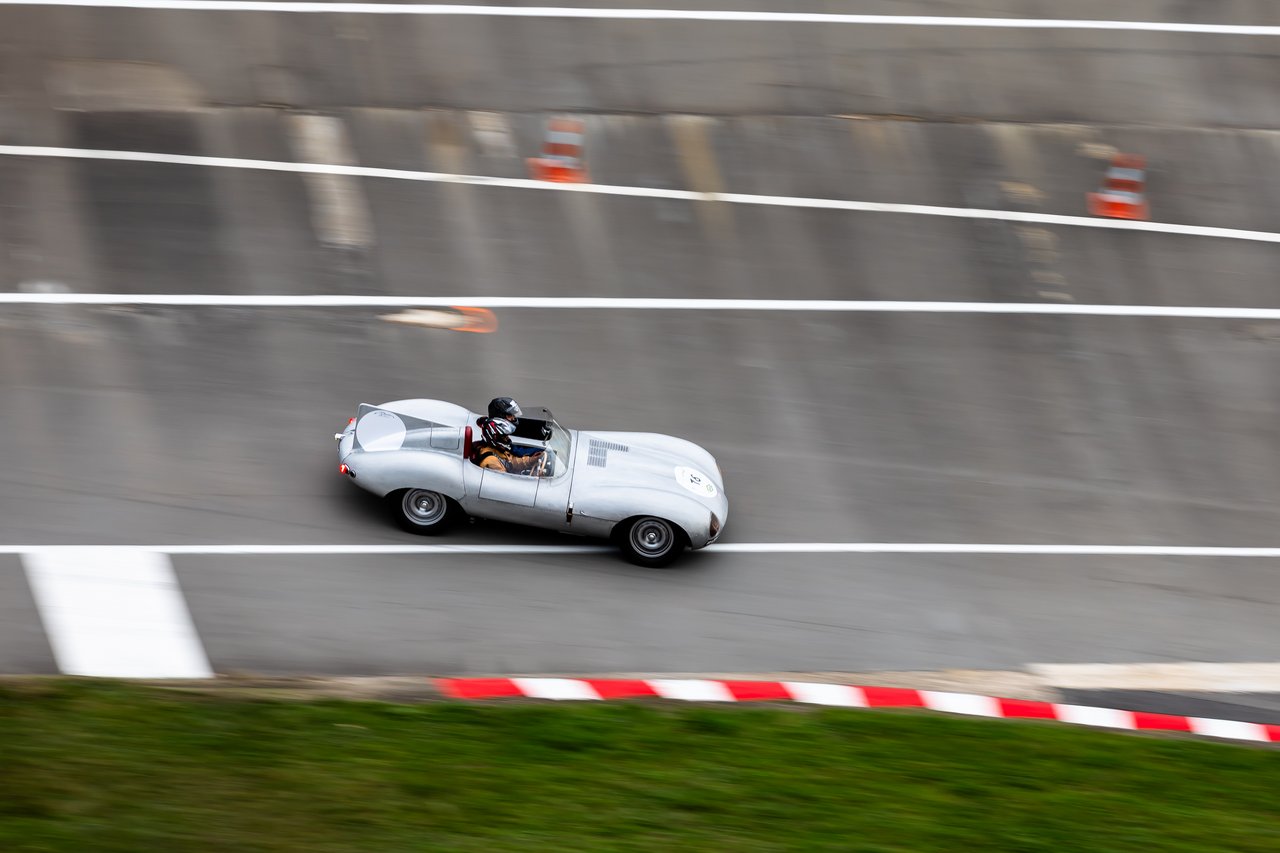 A vintage, light-colored racing car, possibly a classic sports car, is speeding on a racetrack. The background is blurred to convey motion, and red and white striped curbs are visible at the bottom.