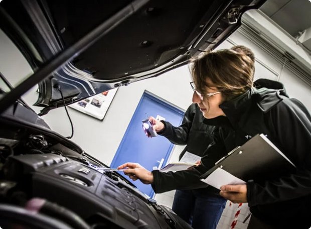 Two people lean over an open car engine bay. One person with glasses and a clipboard is making notes or inspecting something, while the other person might be illuminating or pointing at something.