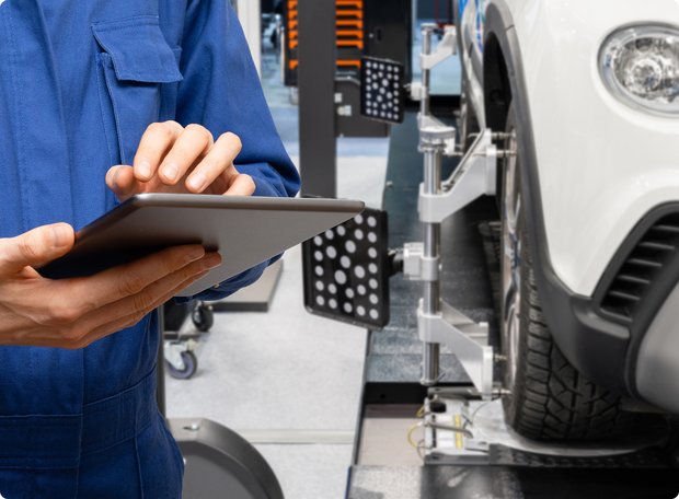 Technician in blue workwear using a tablet during a vehicle alignment in a workshop.