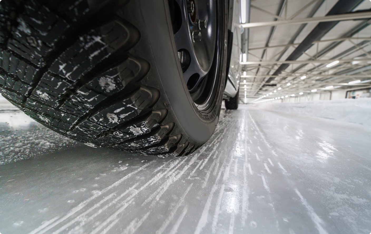 A close-up, low-angle shot of a car tire on a slick, icy or snowy surface, with small ice particles scattered around. The tire tread is visible, and in the blurred background, a large indoor space with a structured ceiling can be seen, suggesting an indoor ice track or testing facility.