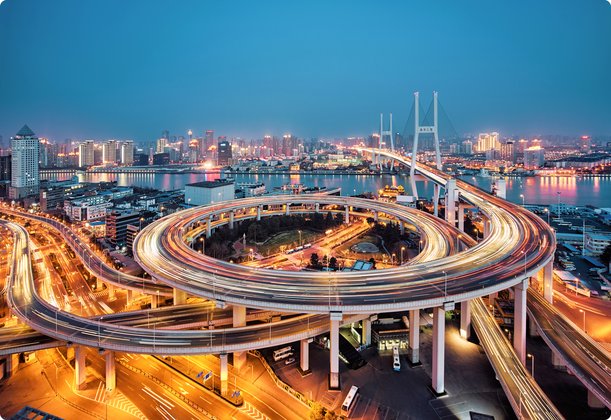 Illuminated highway interchange at night in a modern city with a river and skyscrapers in the background.