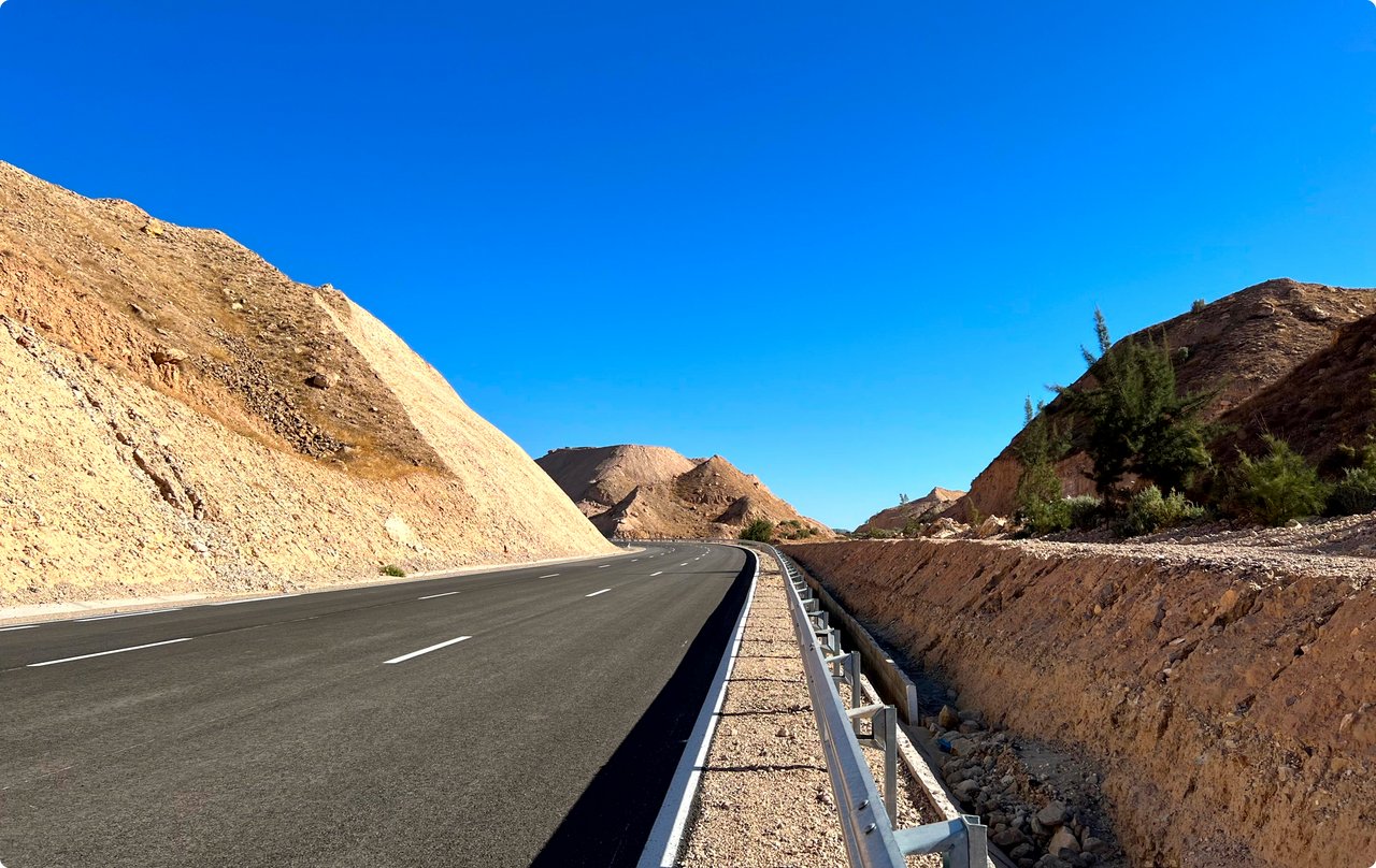 An expansive construction site or newly laid road running through a hilly, earthy landscape. On the left is a freshly graveled or asphalted area with markings, on the right a drainage system and heaped earth mounds under a bright blue sky.