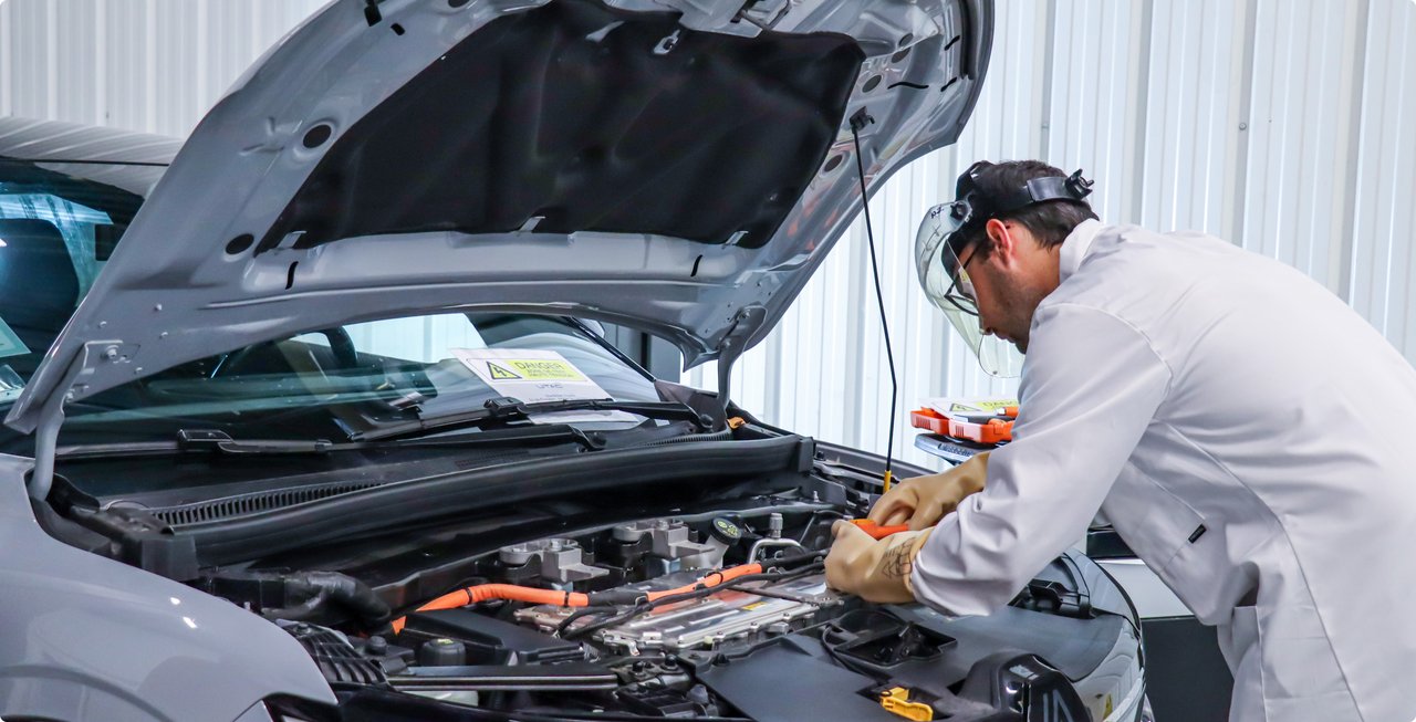 A mechanic or technician in a white lab coat is working on the open engine bay of a car. He is leaning over the engine, which shows many cables and components, and appears to be performing a repair or maintenance.