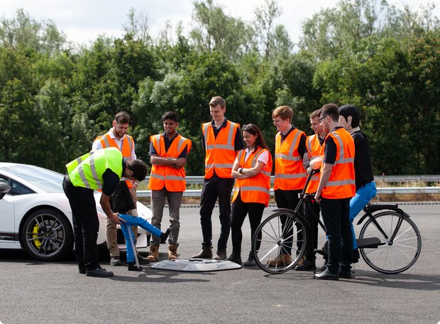A group of people wearing safety vests gathered around a spot on the ground, next to a white sports car and with trees in the background. It appears to be an event or exercise.