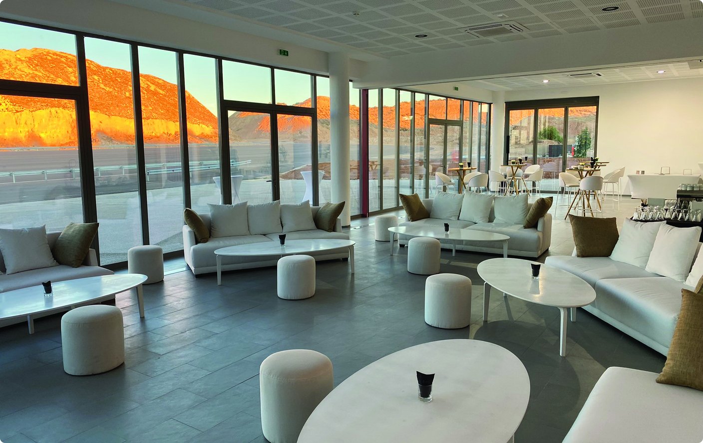 Ein moderner, geräumiger Loungebereich mit zahlreichen weißen Sofas, Couchtischen und Hockern, ausgestattet mit großen Fenstern, die einen Blick auf eine Landschaft mit Hügeln bieten, die von warmem Sonnenlicht, möglicherweise bei Sonnenuntergang oder Sonnenaufgang, beleuchtet werden.