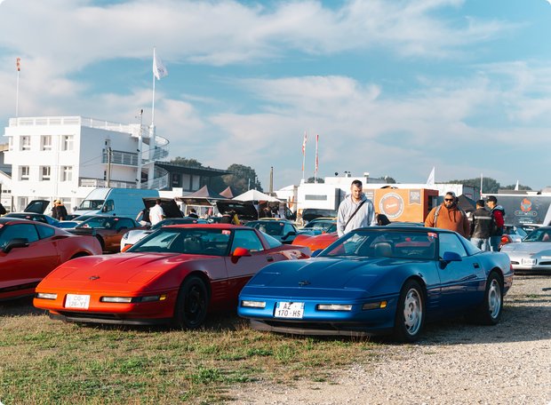 A collection of classic sports cars, including a prominent blue and a red Chevrolet Corvette C4, are parked on a gravel and grass lot during an outdoor event. In the background, white buildings and various people are visible under a partly cloudy sky.