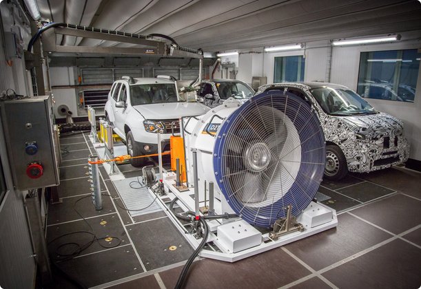 Two cars, one white and the other with a camouflage pattern, are in a testing laboratory equipped with large fans and measurement devices.