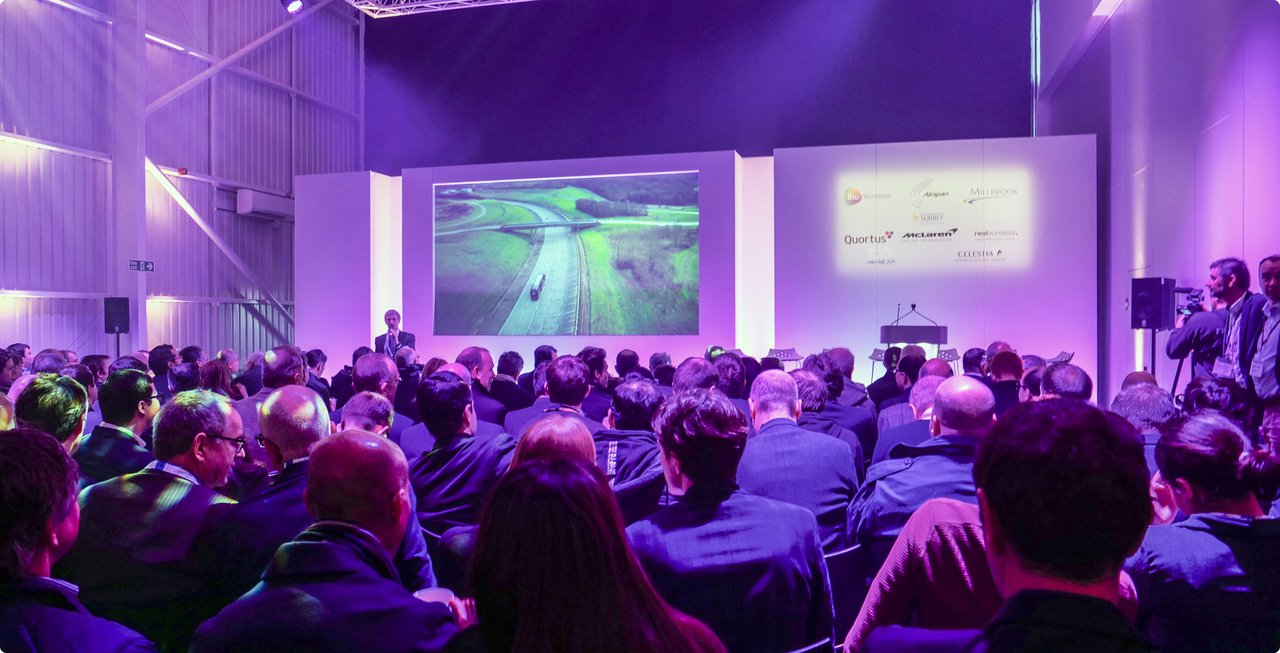 A large group of people are seated in a dimly lit room, facing a stage with two large screens displaying presentations. The room is bathed in purple light.