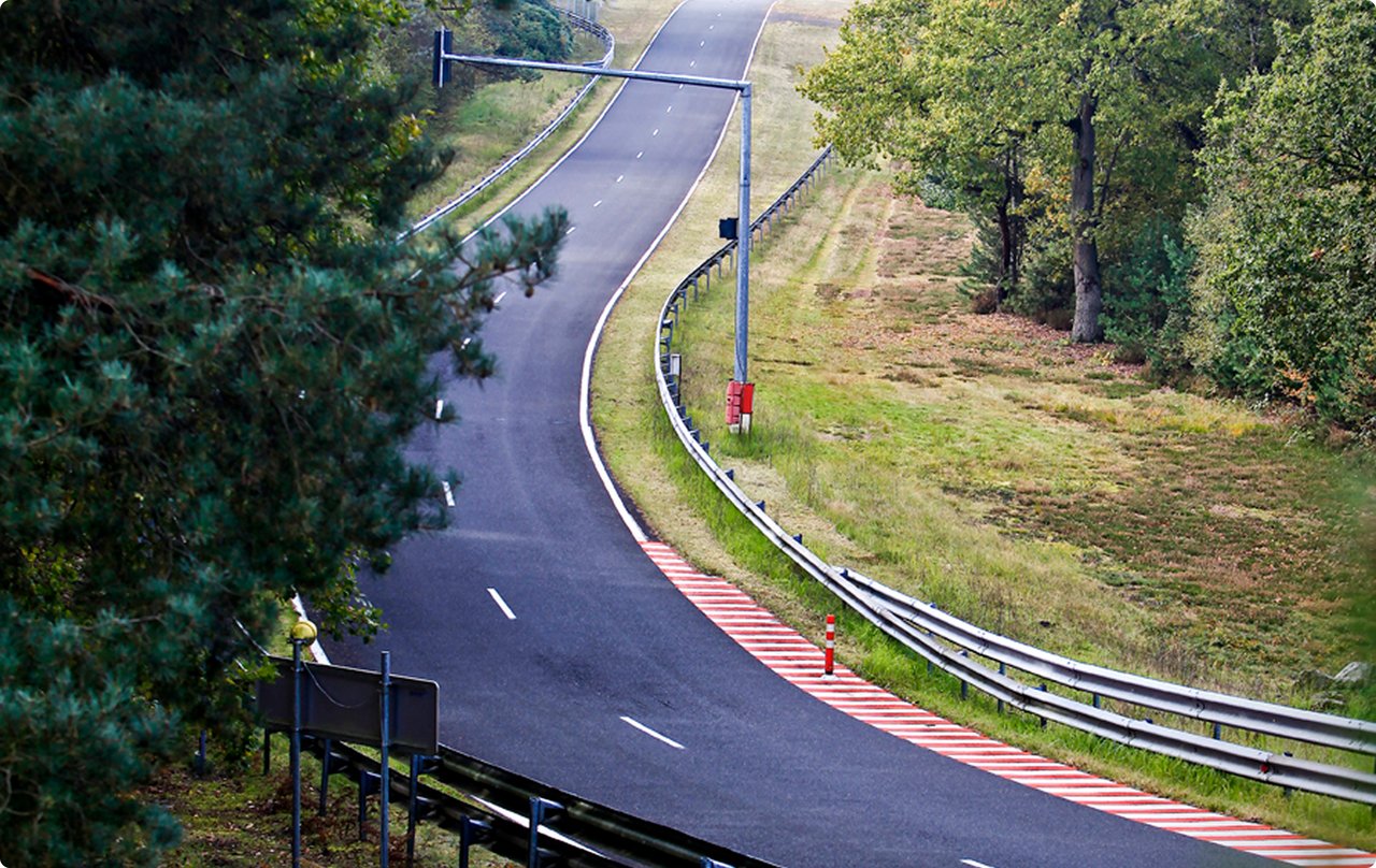 A winding race track with red and white curbs, surrounded by green trees and grass.