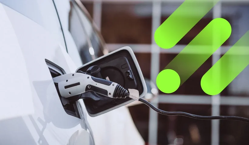 Close-up of a charging station plugged into a white electric car, actively charging it. The charging cable and port are clearly visible. In the upper right area of the image, there are three stylized green shapes, which could symbolize energy or progress.
