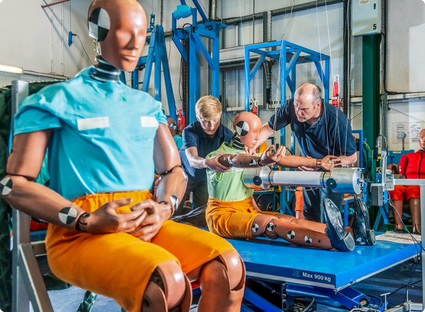 A crash test dummy wearing orange pants and a blue top sits in the foreground of a lab or testing environment, while in the background, two more dummies and technicians are seen preparing for an impact test.