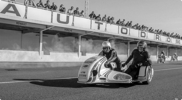 Une image historique en noir et blanc d'une moto side-car sur une piste de course, avec des spectateurs dans une tribune en arrière-plan et le mot "AUTODROM" visible.