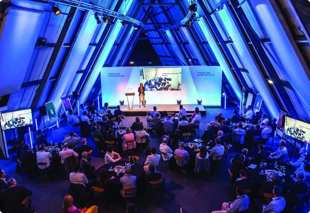 Eine Konferenz oder Veranstaltung in einem großen Saal mit dreieckiger Deckenstruktur, beleuchtet in Blautönen. Eine Person spricht auf einer Bühne mit einer großen Leinwand im Hintergrund, vor einem Publikum, das an Tischen sitzt.