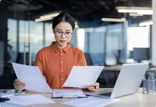 A focused woman with glasses and an orange shirt sits at a desk in a modern office, carefully comparing two documents with a laptop beside her.