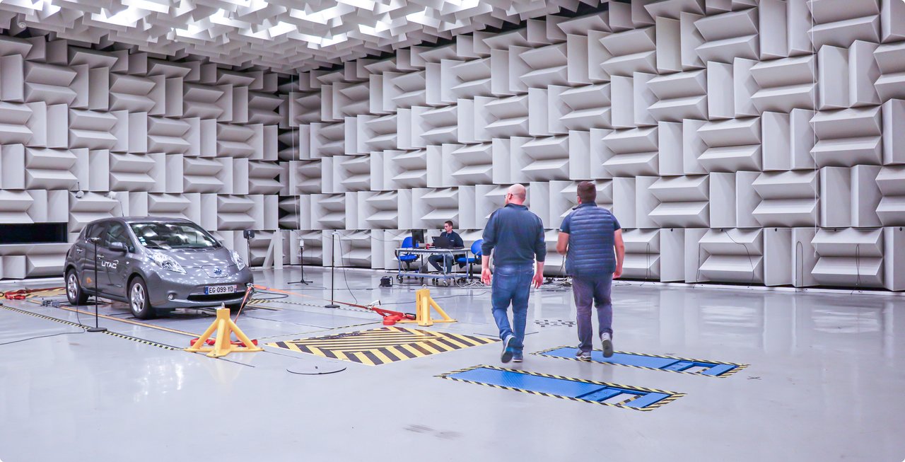 Interior view of an anechoic chamber with a grey car and two technicians conducting measurements. The walls of the room are covered with grey, sound-absorbing wedges.