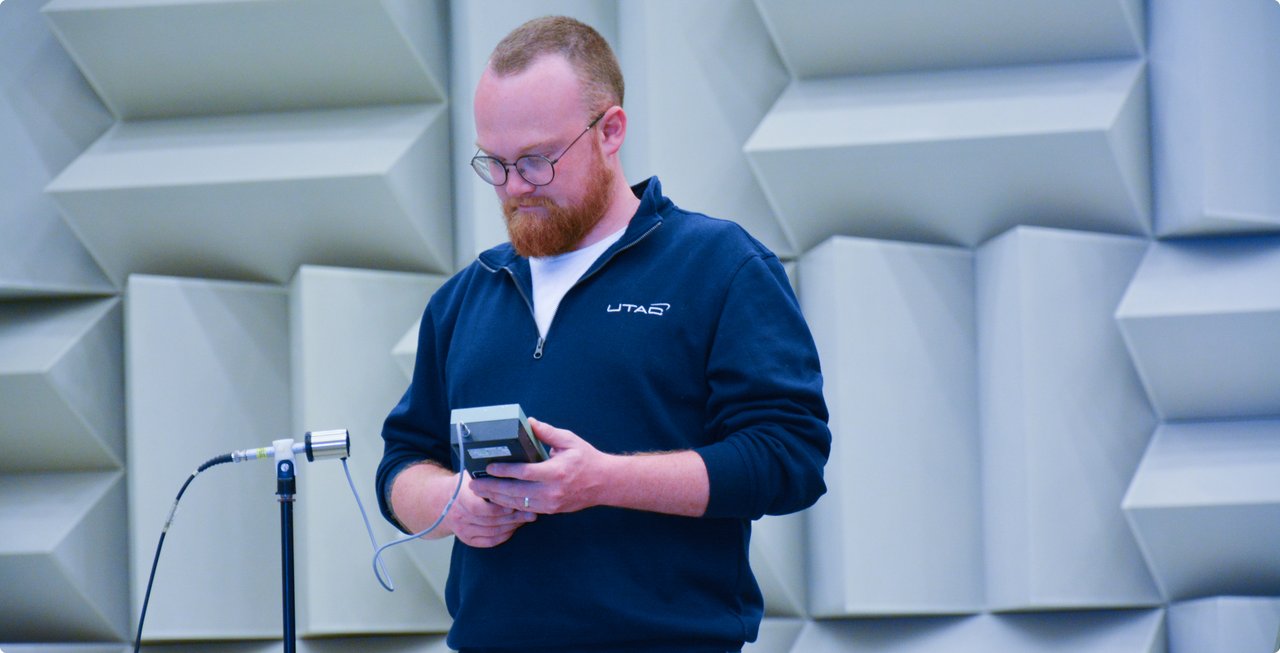 A bearded man with glasses wearing a dark blue sweater, holding a small electronic device in his hands and looking at it intently. He is standing in a room with sound-absorbing walls.