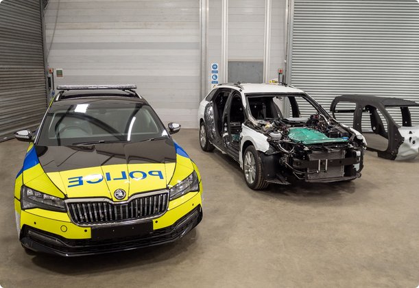 A police vehicle, resembling a Skoda, with yellow and blue livery and a light bar on the roof, stands in the foreground of a hall. Behind it, a severely damaged vehicle missing body panels and a standalone car frame are visible, all appearing to be in a workshop or testing environment for evaluation or repair.
