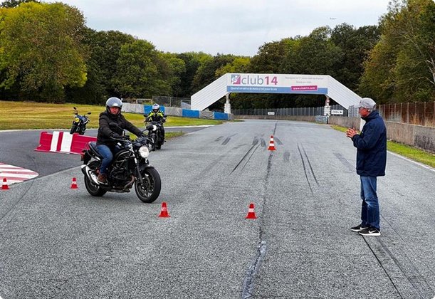 A group of motorcyclists are undergoing riding training on a gravel track marked with cones. A trainer or instructor stands on the side giving instructions. In the background, a finish arch and trees are visible.