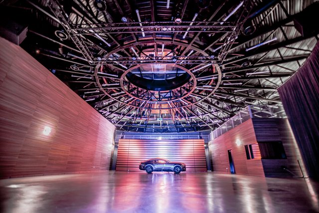 A sleek, dark-colored car is dramatically illuminated in the center of a large, high-ceilinged hall with complex lighting rigs and scaffolding. The walls are adorned with unique, wavy patterns, creating an artistic and futuristic exhibition space.