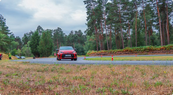 Une voiture rouge roule sur un chemin pavé à travers une zone boisée, jalonnée de cônes orange, sous un ciel nuageux.