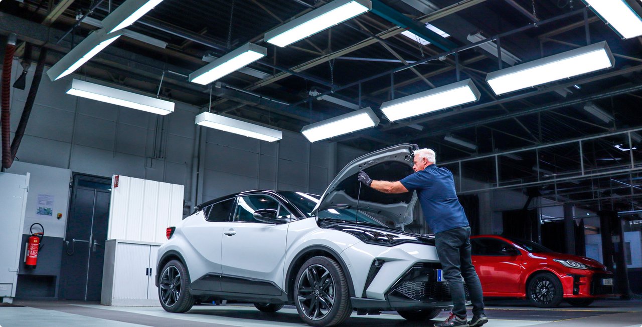 A mechanic leans over the open engine bay of a white car in a brightly lit workshop, with a red vehicle visible in the background.