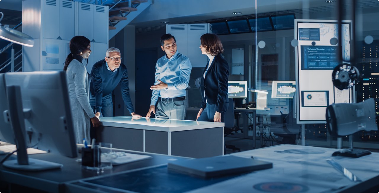 A group of five people are discussing in a modern control room or laboratory around an interactive table with screens in the background.