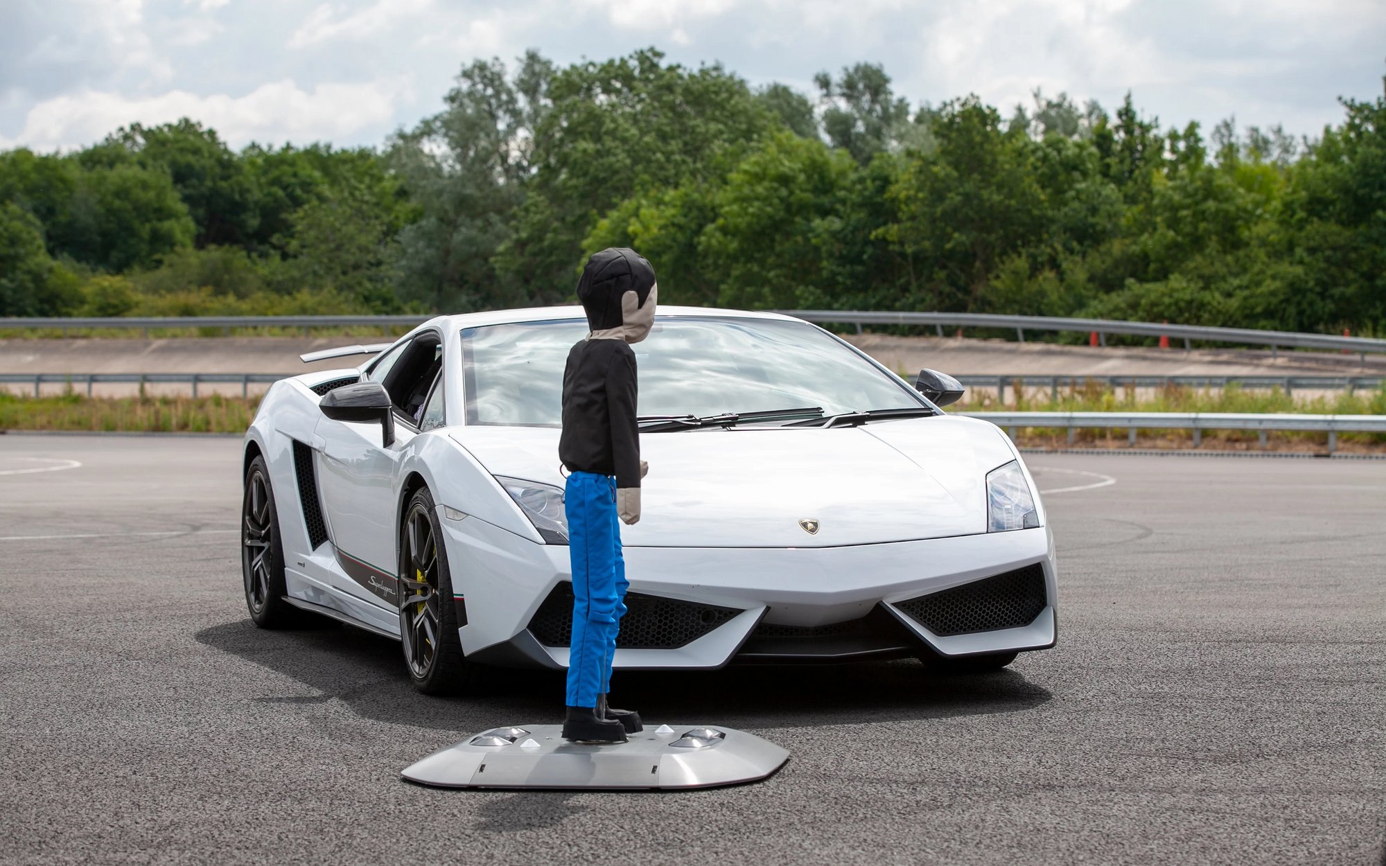 A crash test dummy stands in front of a white sports car coupe on a gravel parking lot, with trees visible in the background.