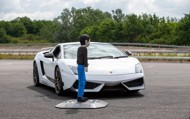 A crash test dummy stands in front of a white sports car coupe on a gravel parking lot, with trees visible in the background.