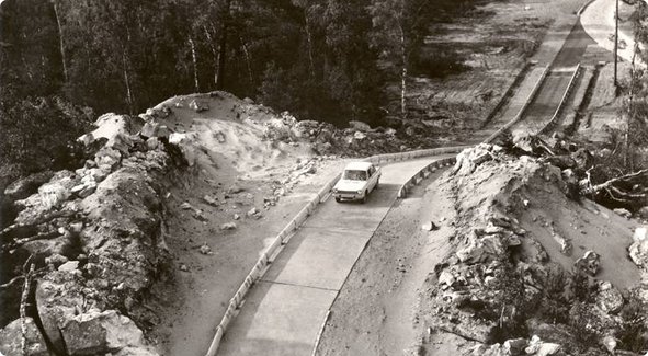 Une image historique en noir et blanc montrant une petite voiture sur une route sinueuse et étroite à travers un paysage vallonné, éventuellement rocheux, avec des arbres en arrière-plan.