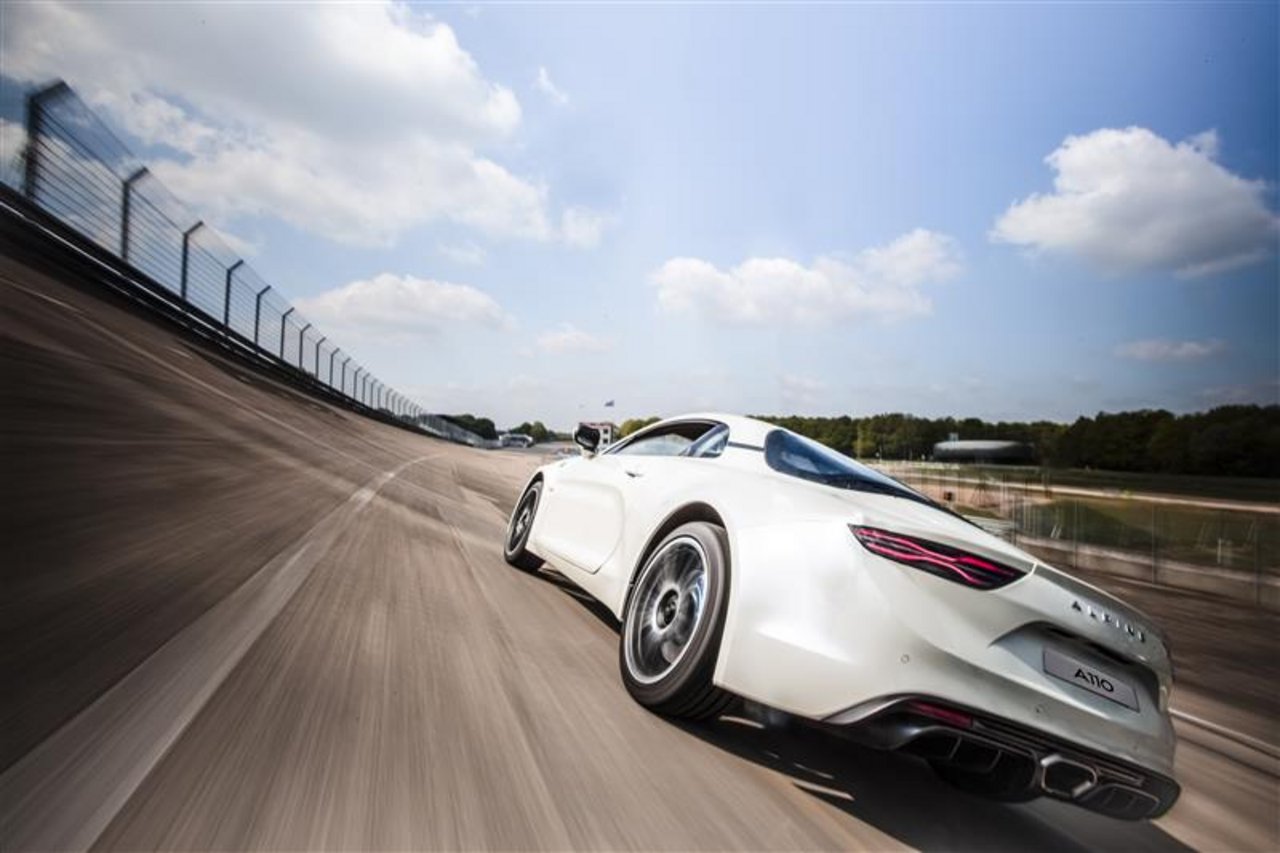 A sleek, futuristic white concept car is driving at an angle on a race track with a high-banked turn visible on the left. The sky is partly cloudy and bright.