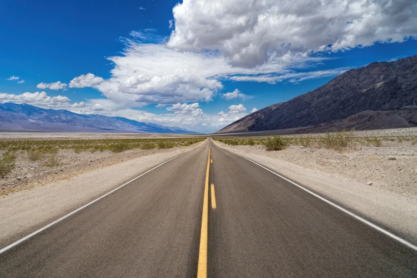 A long, straight asphalt road with a yellow centerline extending through a barren desert or semi-desert landscape. Rocky mountains rise to the left and right of the road, and above them stretches a blue sky with white clouds. The road's perspective leads directly into the distance.