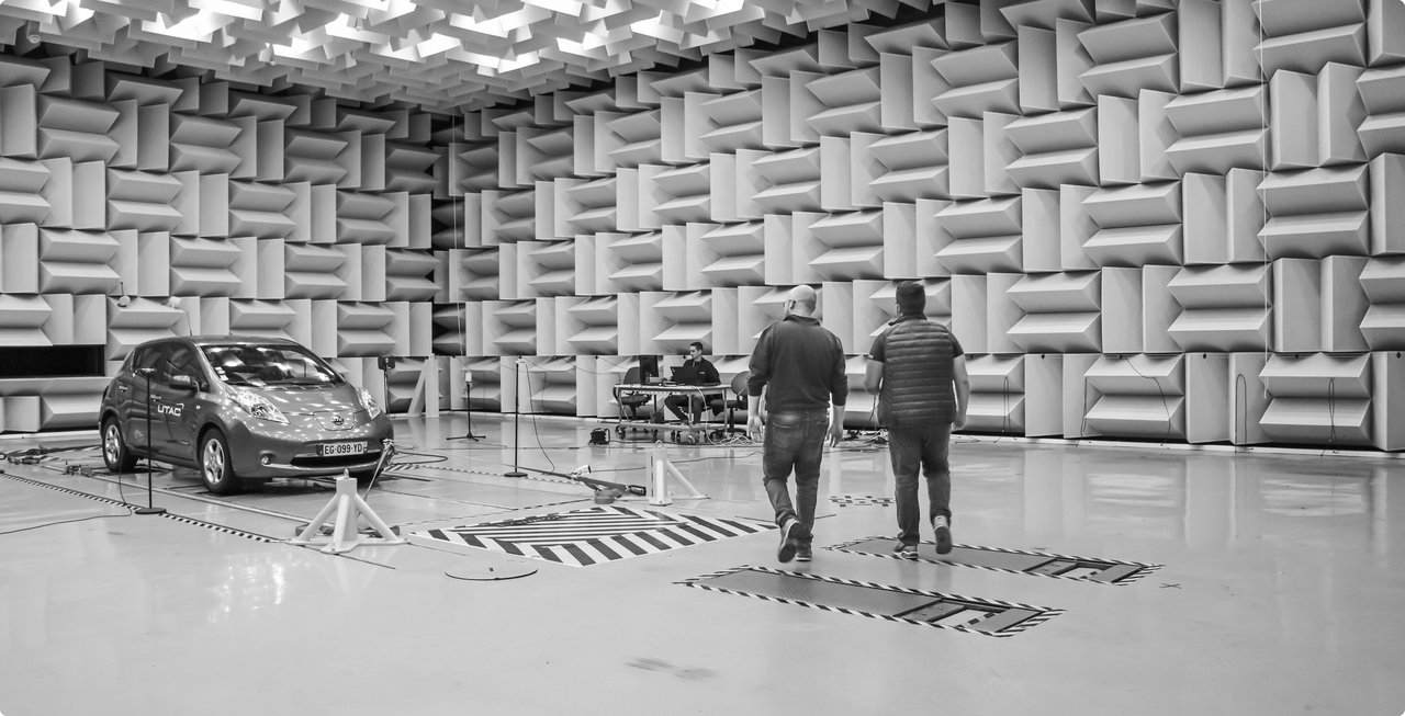 A black and white image of a large anechoic chamber with sound-absorbing wedges on the walls. A car is positioned in the center of the room, and two people stand in the background observing it.