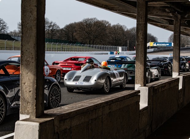 Several classic and modern sports cars are lined up under a covered pit lane or shelter, ready for the track. A silver vintage car is prominent in the center.