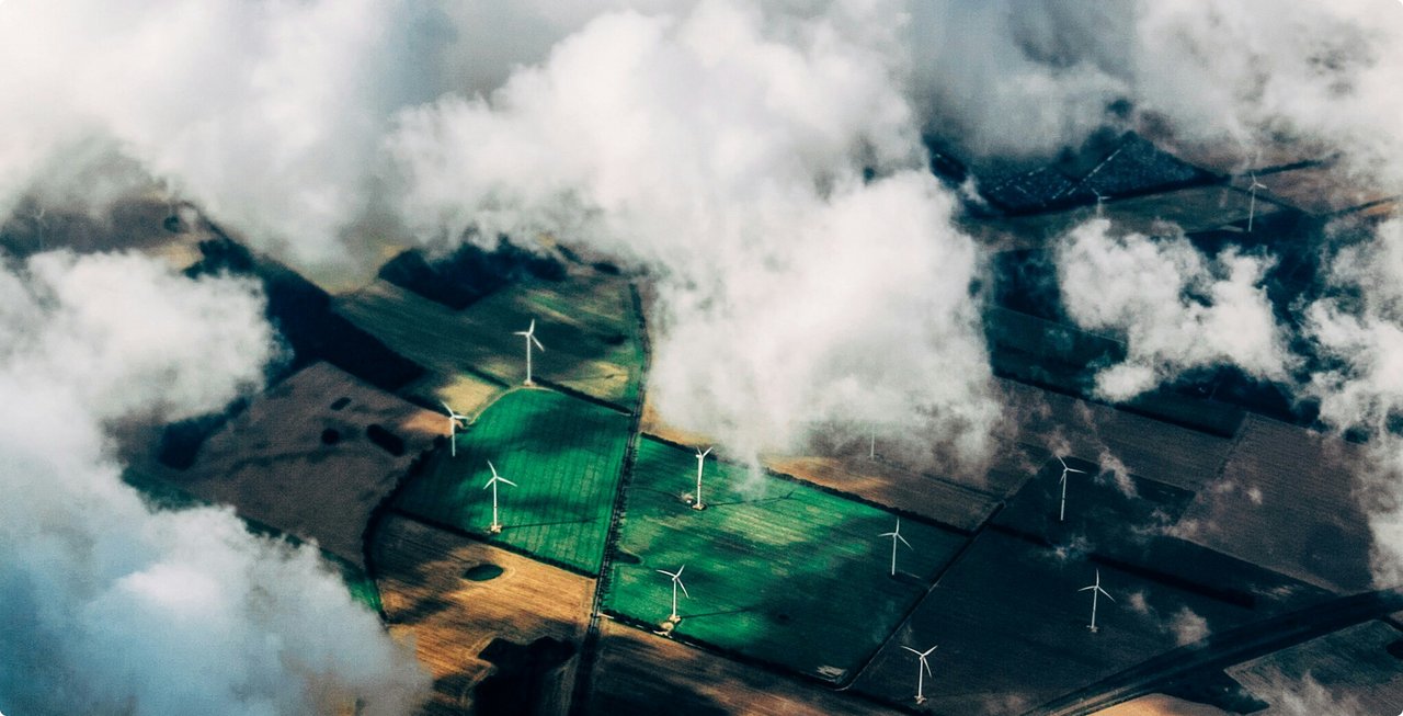 An aerial or satellite view from above, showing a landscape with green fields or bodies of water surrounded by darker areas and clouds. The green areas appear artificially demarcated or cultivated.