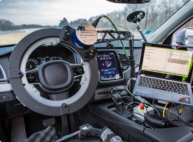 Interior view of a vehicle during a test drive, with extensive measurement equipment installed on the steering wheel and the passenger seat. A laptop displaying technical data is also visible.