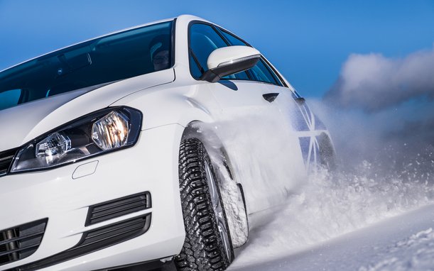 A white car drives dynamically through snow, with snow being kicked up by the tire. The sky is blue.