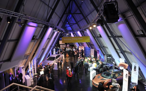 An interior view of a large, modern hall with a high, pointed ceiling structure made of metal struts. The hall is lit for an event, with purple accents and an illuminated display in the distance. Numerous people are scattered across the floor, viewing exhibits, including vehicles and presentation stands.
